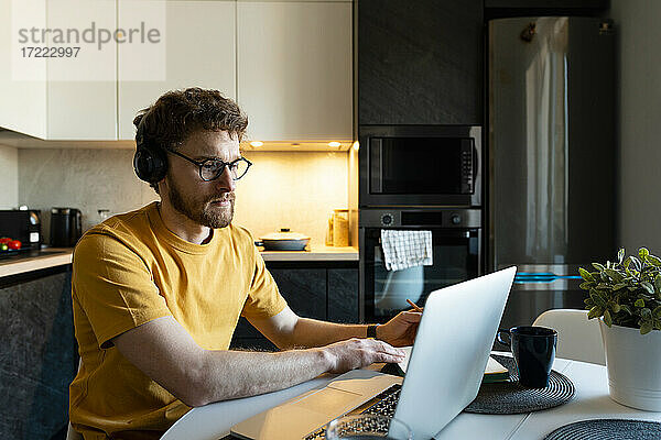 Männlicher Freiberufler hört über Kopfhörer zu  während er in der Küche einen Laptop benutzt