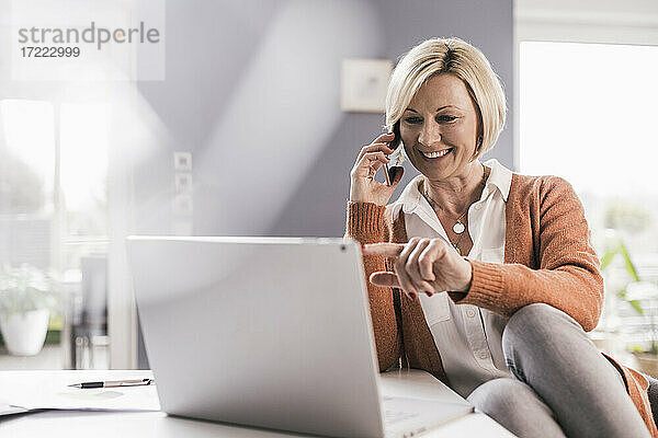 Lächelnde weibliche Fachkraft  die auf einen Laptop zeigt  während sie über ein Smartphone zu Hause kommuniziert