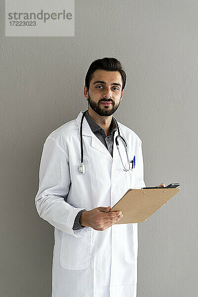 Gut aussehender männlicher Arzt mit Klemmbrett vor einer Wand stehend