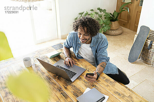 Geschäftsmann  der ein Smartphone hält  während er einen Laptop auf einem Tisch zu Hause benutzt