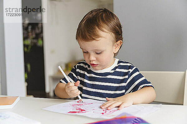 Netter Junge Zeichnung auf Papier mit Bleistift sitzt auf dem Tisch zu Hause