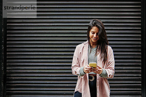Lächelnde Frau  die ein Mobiltelefon benutzt  während sie vor einem Fensterladen steht