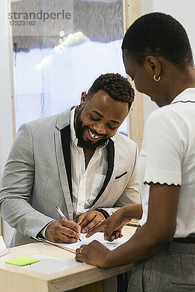 Lächelnder Geschäftsmann  der auf einem Papier schreibt  während er neben einem Kollegen im Büro steht