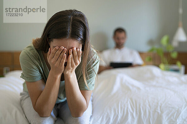 Verzweifelte Frau  die ihr Gesicht bedeckt und auf dem Bett sitzt  während ihr Freund im Hintergrund ein digitales Tablet benutzt