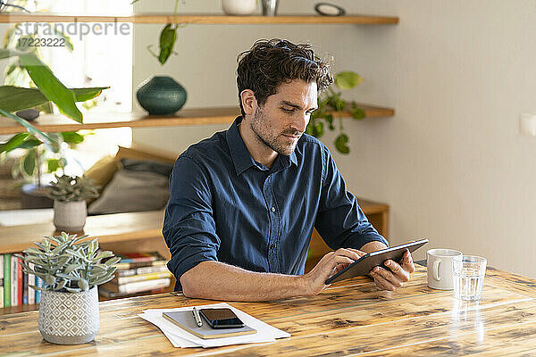 Nachdenklicher männlicher Freiberufler  der ein digitales Tablet benutzt  während er am Tisch im Heimbüro sitzt