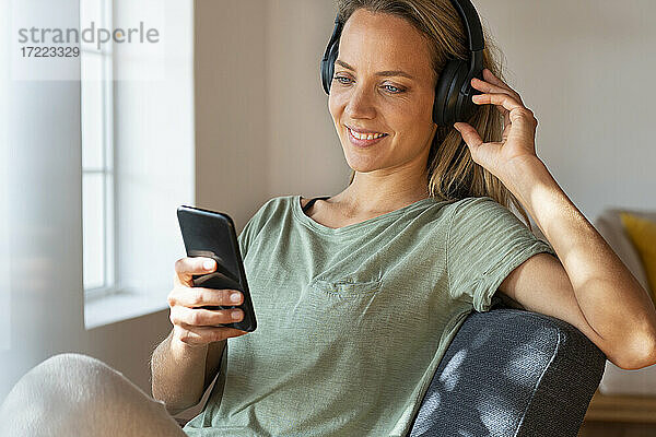 Lächelnde Frau  die über Kopfhörer Musik hört  während sie zu Hause ein Mobiltelefon benutzt