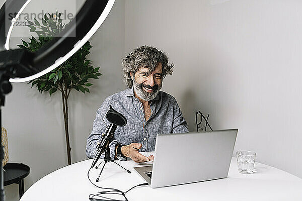Älterer männlicher Freiberufler beim Podcasting mit Laptop auf dem Tisch im Heimbüro