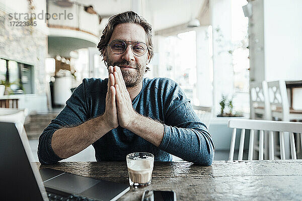 Lächelnder Mann mit verschränkten Händen am Tisch in einem Café