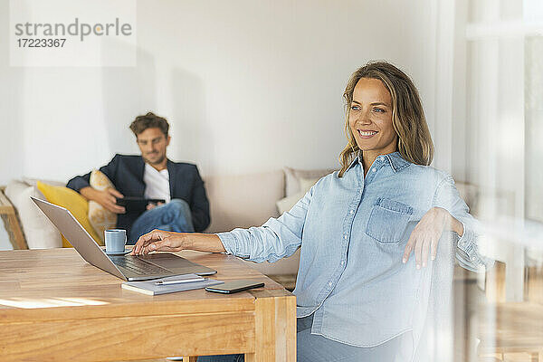 Nachdenkliche Frau mit Laptop am Tisch  während ihr Freund zu Hause im Büro sitzt