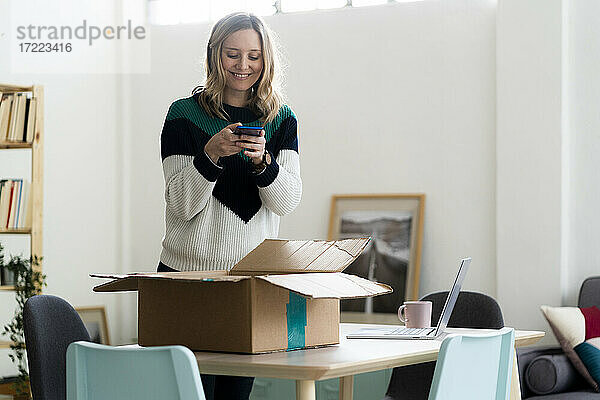 Lächelnde Frau  die ein Foto von einem Paketkarton durch ein Mobiltelefon zu Hause macht