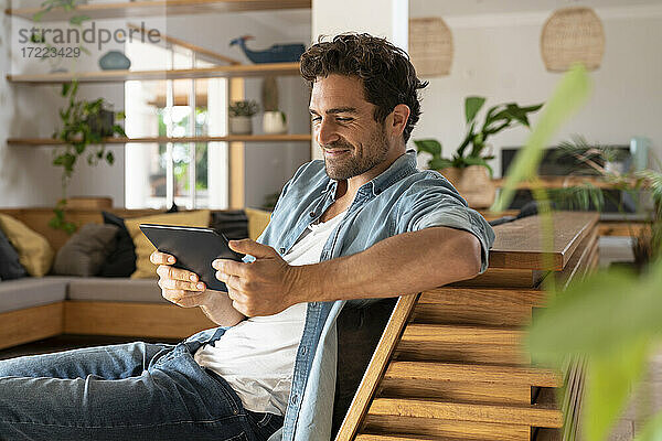 Lächelnder mittelgroßer erwachsener Mann  der auf ein digitales Tablet schaut  während er zu Hause auf der Couch sitzt