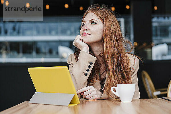 Schöne Frau mit Hand am Kinn träumt  während sie vor einem digitalen Tablet und einer Kaffeetasse im Café sitzt