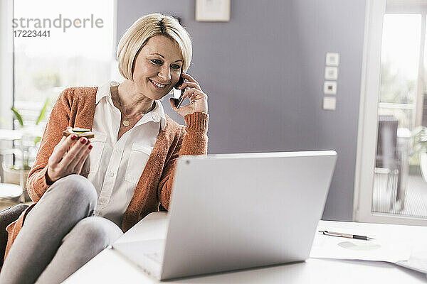 Lächelnde Unternehmerin  die mit ihrem Smartphone telefoniert und etwas isst  während sie zu Hause arbeitet