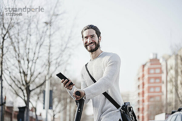 Glücklicher männlicher Hipster  der sein Smartphone auf einem Motorroller hält
