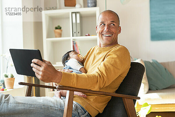 Lächelnder Vater schaut weg  während er seine schlafende Tochter und ein digitales Tablet im Wohnzimmer hält