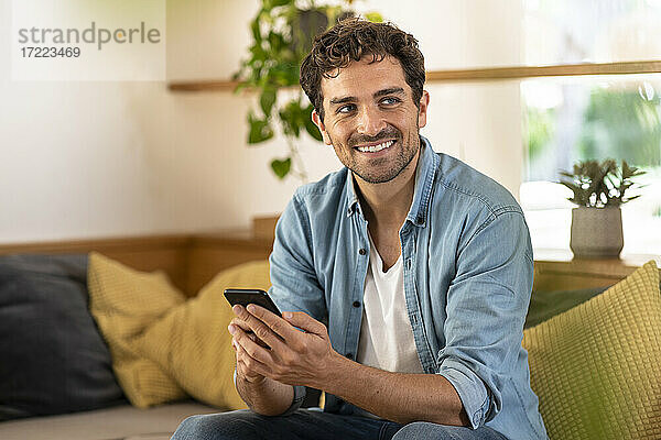 Lächelnder gutaussehender Mann  der sein Smartphone hält  während er auf dem Sofa sitzt