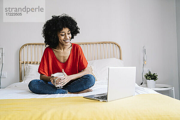 Junge Frau mit Tasse  die auf einen Laptop schaut  während sie zu Hause auf dem Bett sitzt