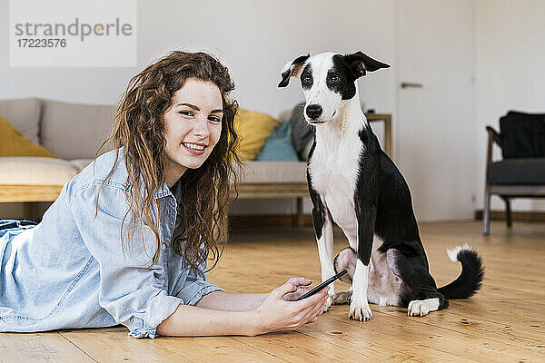 Hübsche Frau liegt auf dem Holzboden und benutzt ein Smartphone  während der Hund neben ihr sitzt und beide in die Kamera schauen