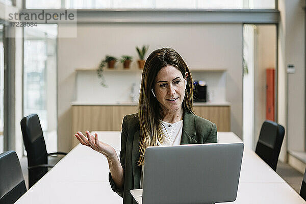 Geschäftsfrau mit In-Ear-Kopfhörern gestikuliert während eines Videogesprächs am Laptop im Büro