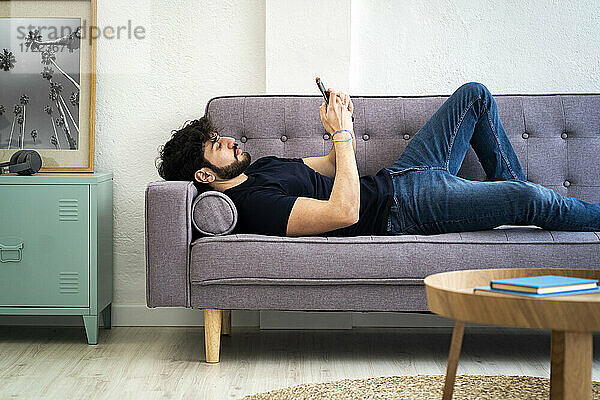 Entspannter junger Mann  der sein Smartphone benutzt  während er zu Hause auf dem Sofa liegt