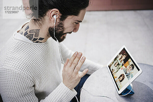 Mann mit Tätowierung winkt mit der Hand während eines Videoanrufs über ein digitales Tablet in einem Straßencafé