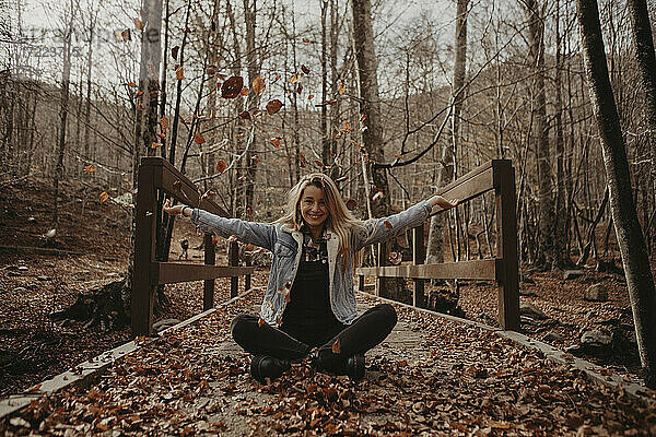Glückliche Frau spielt mit Herbstblättern  während sie auf einer Holzbrücke im Wald sitzt