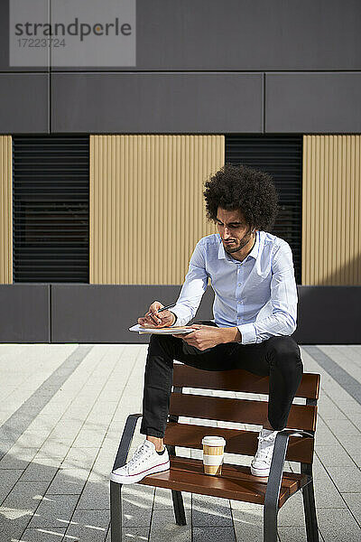 Mann mit Kaffeetasse  der auf einem Stuhl sitzend in ein Buch schreibt  während er an einem sonnigen Tag sitzt