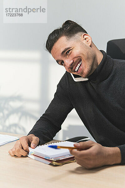 Lächelnder männlicher Finanzberater mit Tagebuch und Stift im Gespräch mit einem Smartphone im Büro
