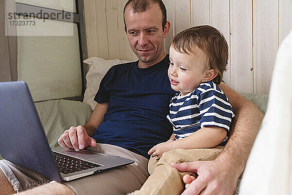 Vater arbeitet am Laptop  während er mit seinem Sohn im Heimbüro sitzt