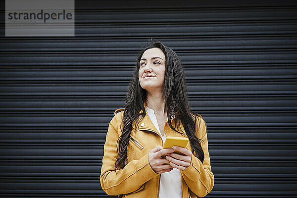 Lächelnde Frau mit Handy schaut weg  während sie vor einem schwarzen Fensterladen steht
