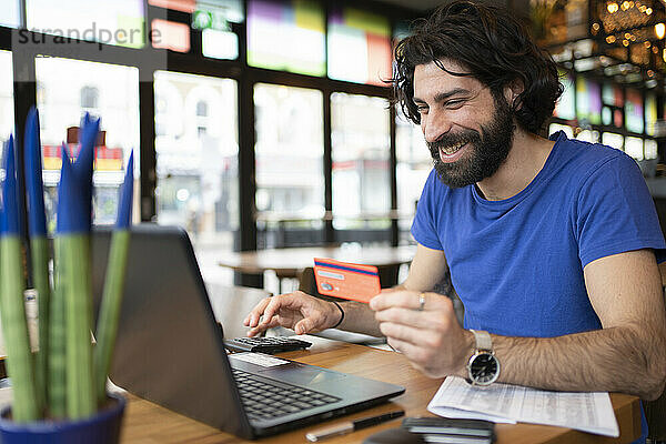Lächelnder Geschäftsmann beim Online-Shopping mit Kreditkarte vor dem Laptop in einem Coworking-Büro