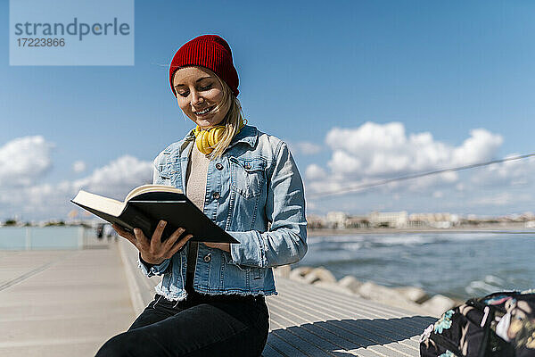 Lächelnde Frau mit Strickmütze liest Tagebuch  während sie auf einer Stützmauer gegen den Himmel sitzt
