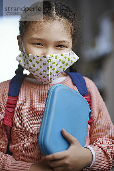 Mädchen mit Gesichtsschutzmaske  das während einer Pandemie ein Mäppchen hält