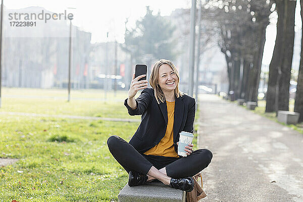 Fröhliche Geschäftsfrau  die ein Selfie mit ihrem Mobiltelefon macht  während sie auf einer Bank im Park sitzt