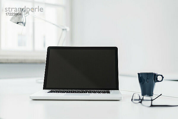 Laptop neben Kaffeetasse und Brille auf dem Schreibtisch im Büro