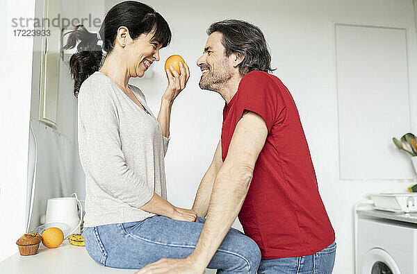 Frau hält eine Orange und sieht einen Mann an  während sie zu Hause auf dem Küchentisch sitzt
