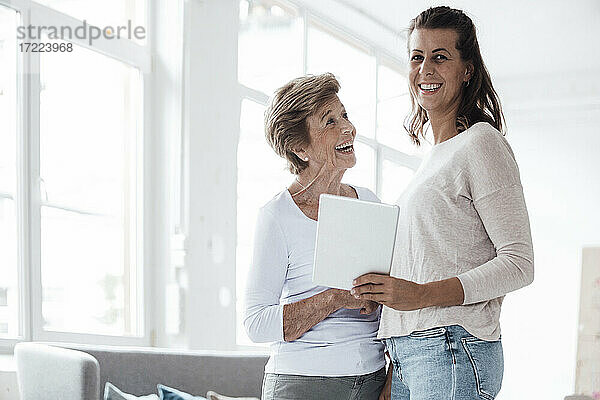 Fröhliche ältere Frau schaut auf ihre Enkelin  die zu Hause ein digitales Tablet hält