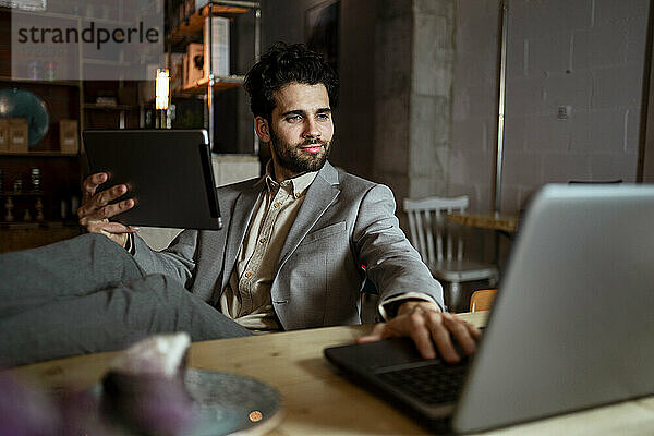 Gutaussehender männlicher Unternehmer mit digitalem Tablet  der in einem Café an einem Laptop arbeitet