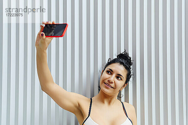 Lächelnde Sportlerin  die ein Selfie mit ihrem Handy vor einer gestreiften Wand macht