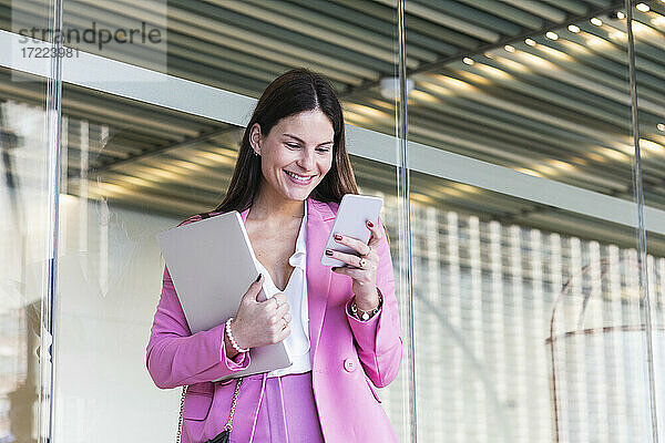 Lächelnde Geschäftsfrau  die ein Mobiltelefon benutzt  während sie mit einem Laptop vor einer Glaswand steht