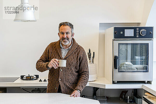 Lächelnder Mann mit Kaffeetasse in der Küche zu Hause stehend