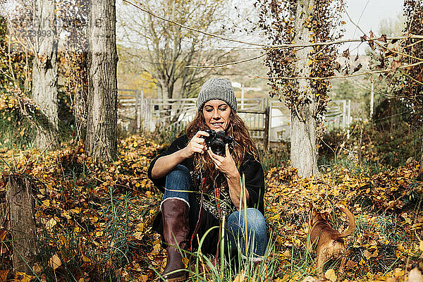 Lächelnde Frau  die durch die Kamera fotografiert  während sie mit ihrem Hund inmitten von Herbstblättern sitzt