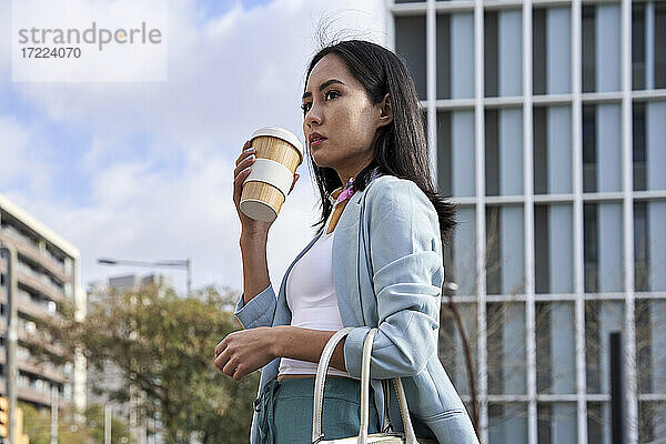 Geschäftsfrau schaut beim Kaffeetrinken vor einem modernen Gebäude weg