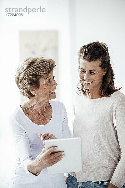 Aufgeregte ältere Frau  die ein digitales Tablet hält  während sie sich mit ihrer Enkelin zu Hause unterhält