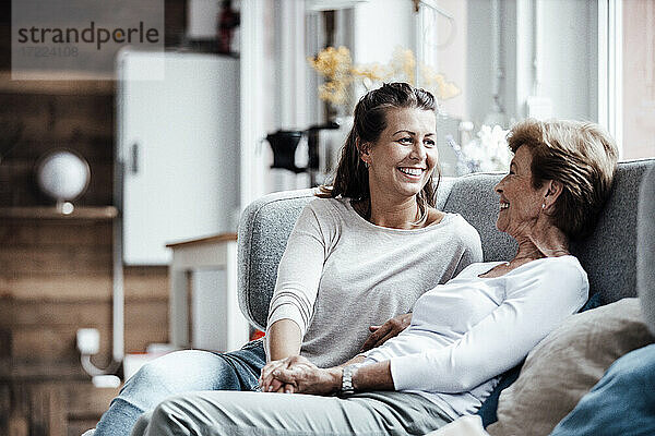 Lächelnde schöne Frau  die die Hand ihrer Großmutter hält  während sie auf dem Sofa zu Hause sitzt