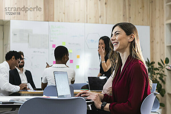 Fröhlicher Unternehmer am Laptop sitzend mit Kollegen im Hintergrund im Büro