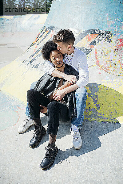 Junger Mann küsst seine Freundin auf die Stirn  während er in einem Skateboard-Park an einem sonnigen Tag sitzt