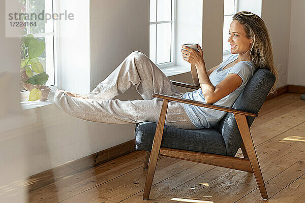 Glückliche Frau hält Kaffeetasse  während sie am Fenster im Wohnzimmer sitzt