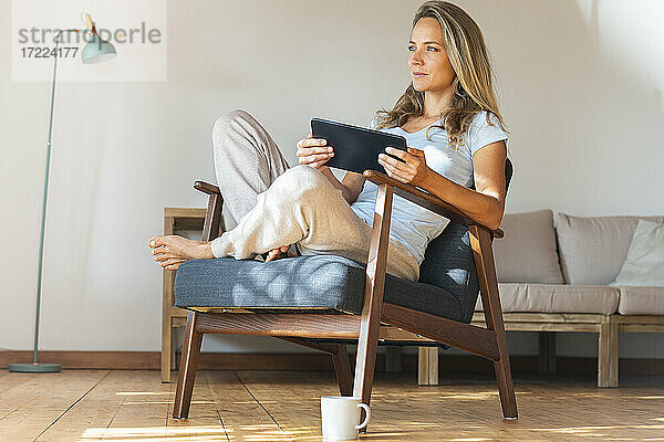 Nachdenkliche Frau  die ein digitales Tablet hält  während sie auf einem Sessel im Wohnzimmer sitzt