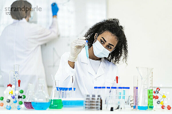 Eine Forscherin untersucht eine Flüssigkeit in einem Reagenzglas  während ein Kollege im Hintergrund im Labor arbeitet